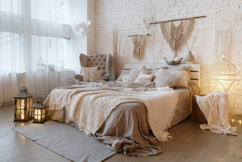 Bočný pohľad na krásnu spálňu s plédom a vankúšmi na pohodlnej posteli, domácou výzdobou a vankúšmi na mäkkom kresle v bielom interiérovom dizajne v bohémskom štýle