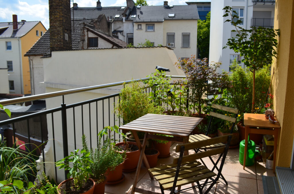 Rastliny, stoly a stoličky na balkóne v jarný / letný deň so starou architektúrou v pozadí.